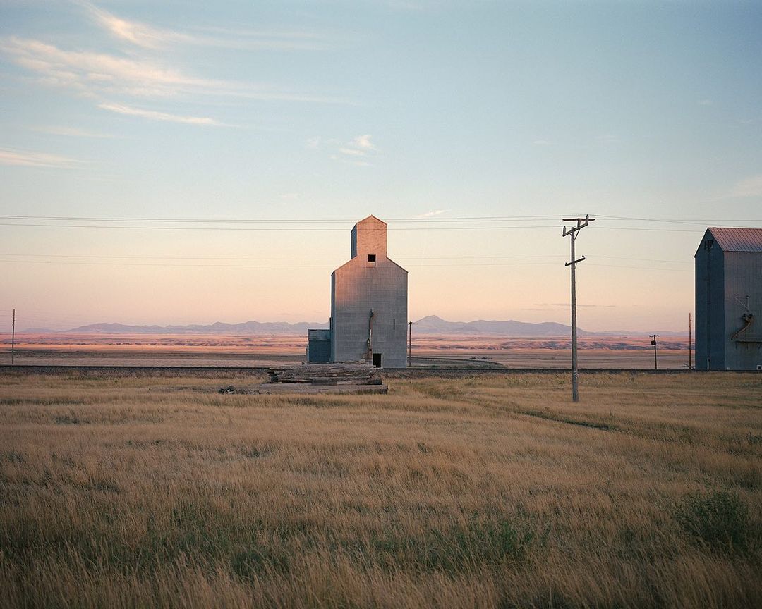 Grain elevator stands in a field with railroad tracks and mountains in the distance under a blue and pink sky at sunset.