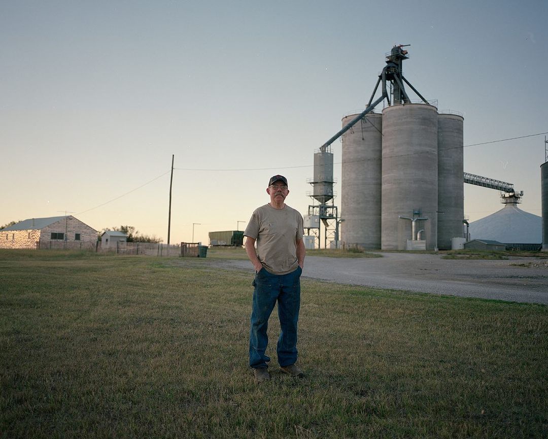 Man stands in a field in front of grain silos at sunset, wearing a baseball cap and jeans, with rural buildings in background.