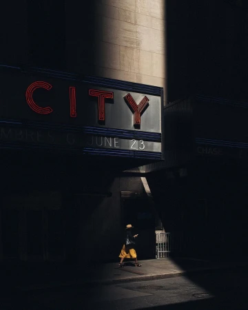 A person in a yellow skirt and hat walks beneath a glowing red neon sign reading CITY on a dark city street.