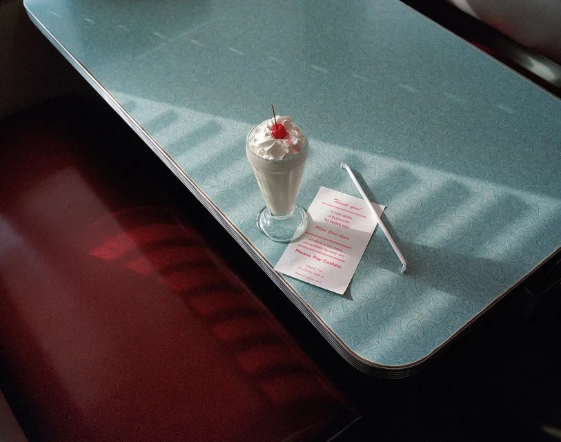 Milkshake with whipped cream and cherry on a diner table, accompanied by a straw and a thank you note in a vintage setting.