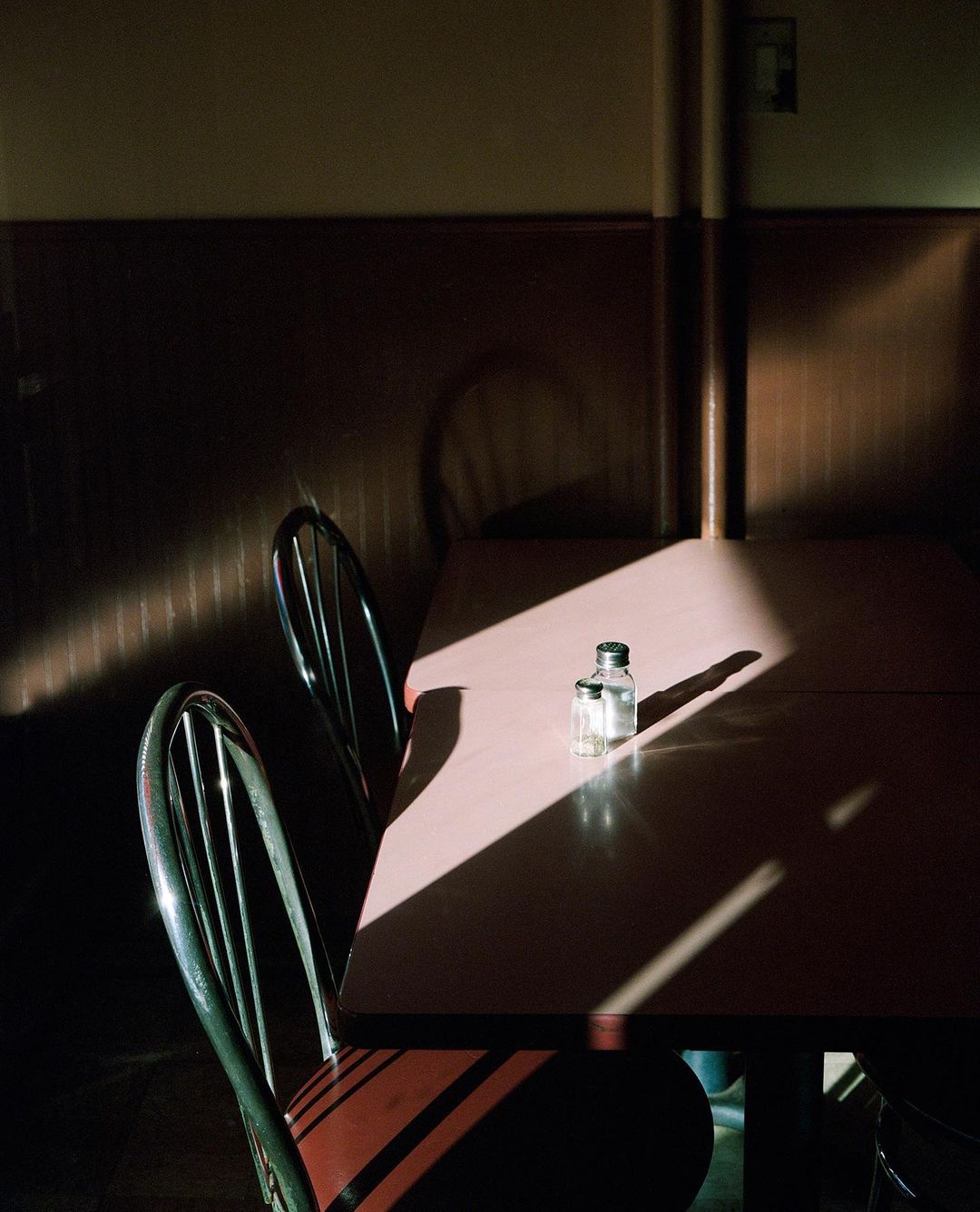 Still life photograph of a glass salt shaker on a table with chairs in a dimly lit room with a warm color palette and film grain.