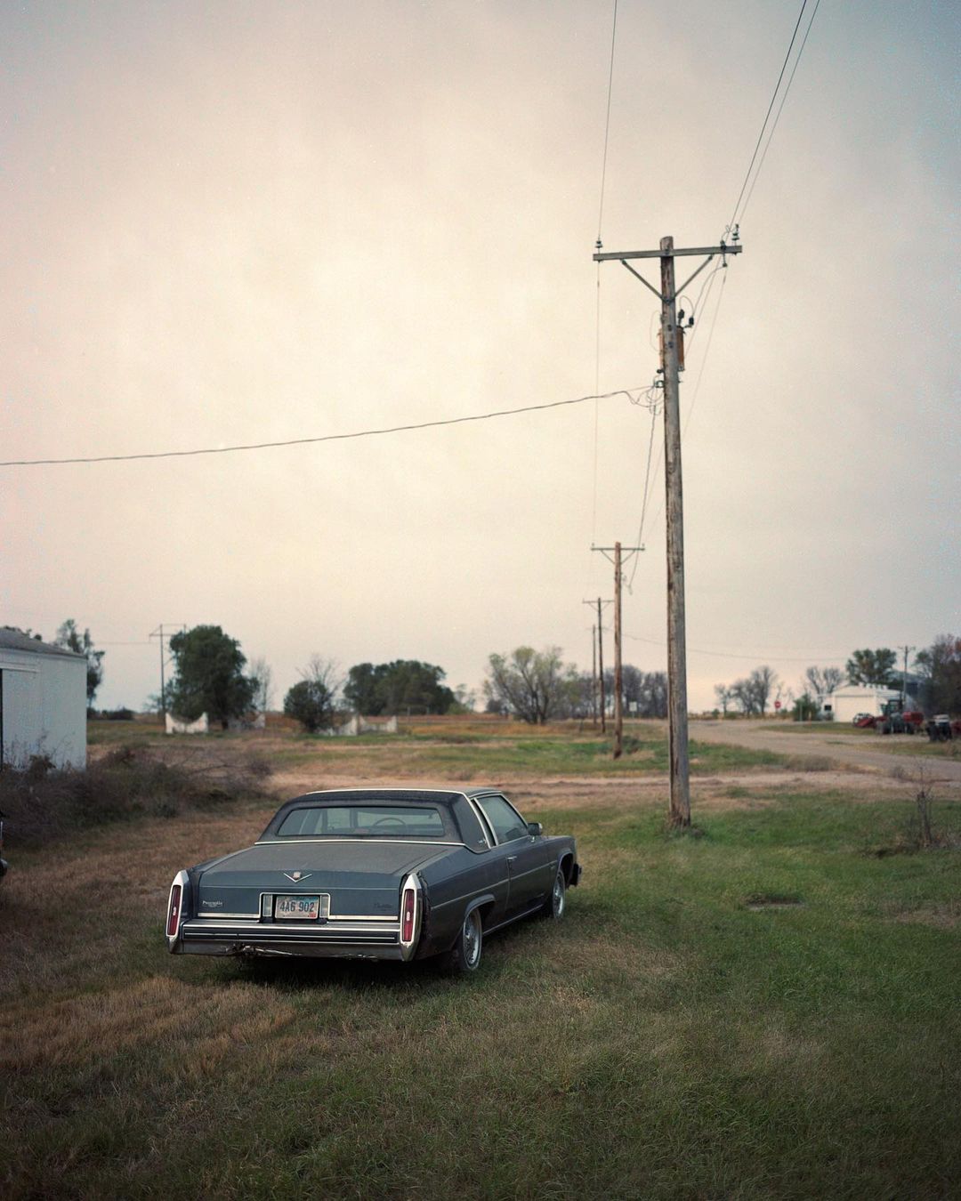 Vintage Cadillac parked on a grassy field with telephone poles receding into the distance under an overcast sky in a rural setting.