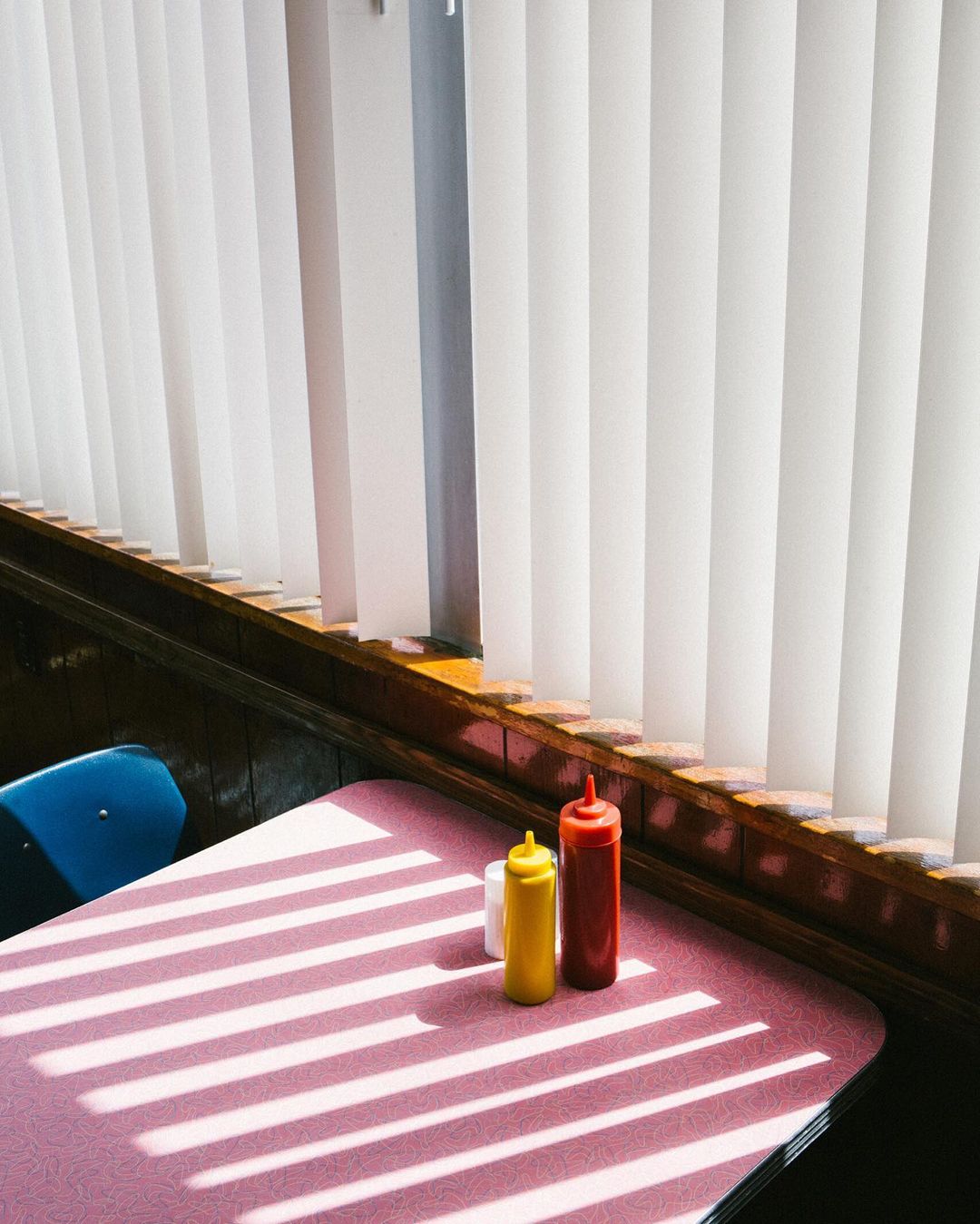 Still life of mustard, ketchup, and salt shaker on a pink diner table with window blinds casting shadows in an indoor setting.