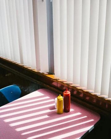 Still life of mustard, ketchup, and salt shaker on a pink diner table with window blinds casting shadows in an indoor setting.