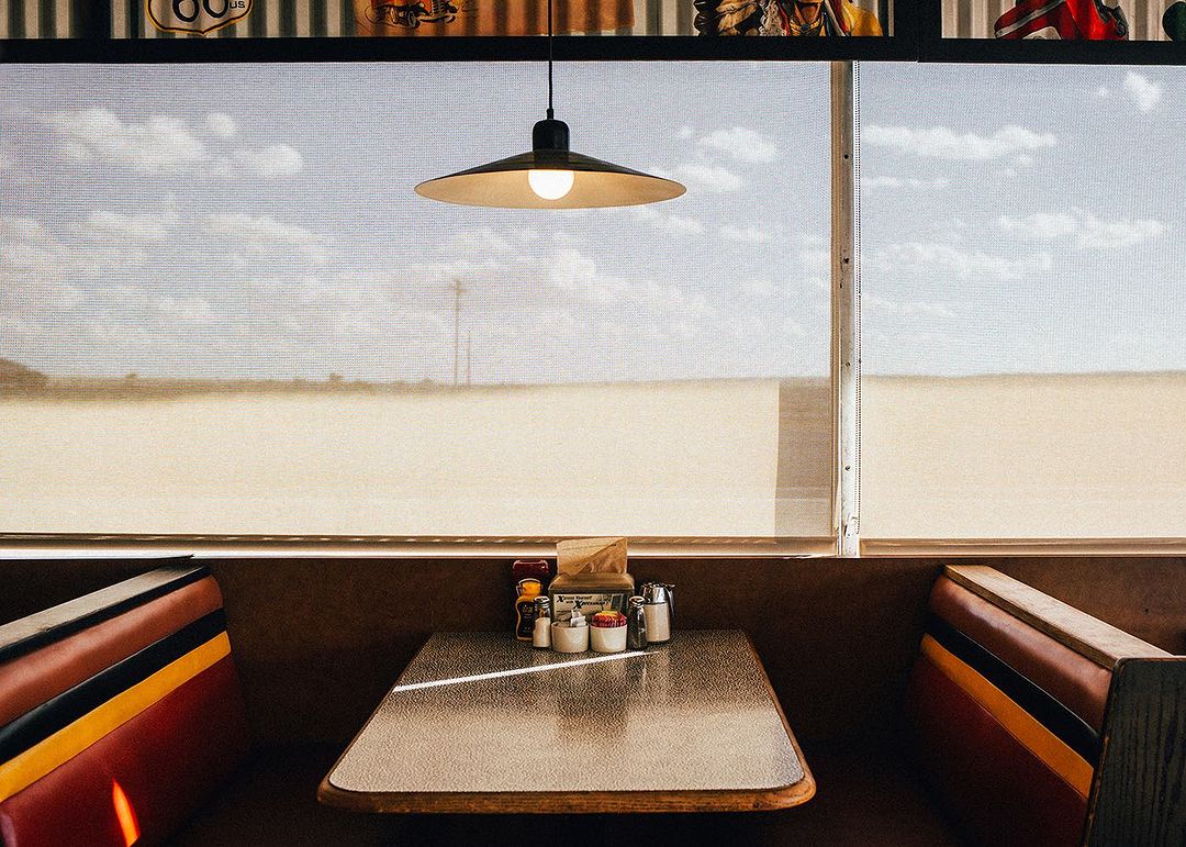 Diner booth with a table set for a meal, window view of a desert landscape, and hanging lamp providing warm illumination.