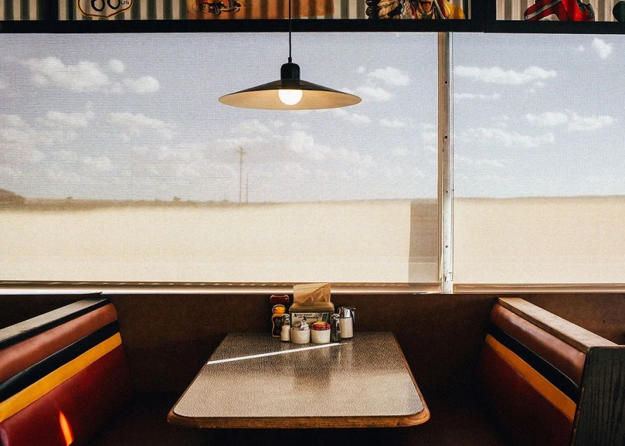 Diner booth with a table set for a meal, window view of a desert landscape, and hanging lamp providing warm illumination.