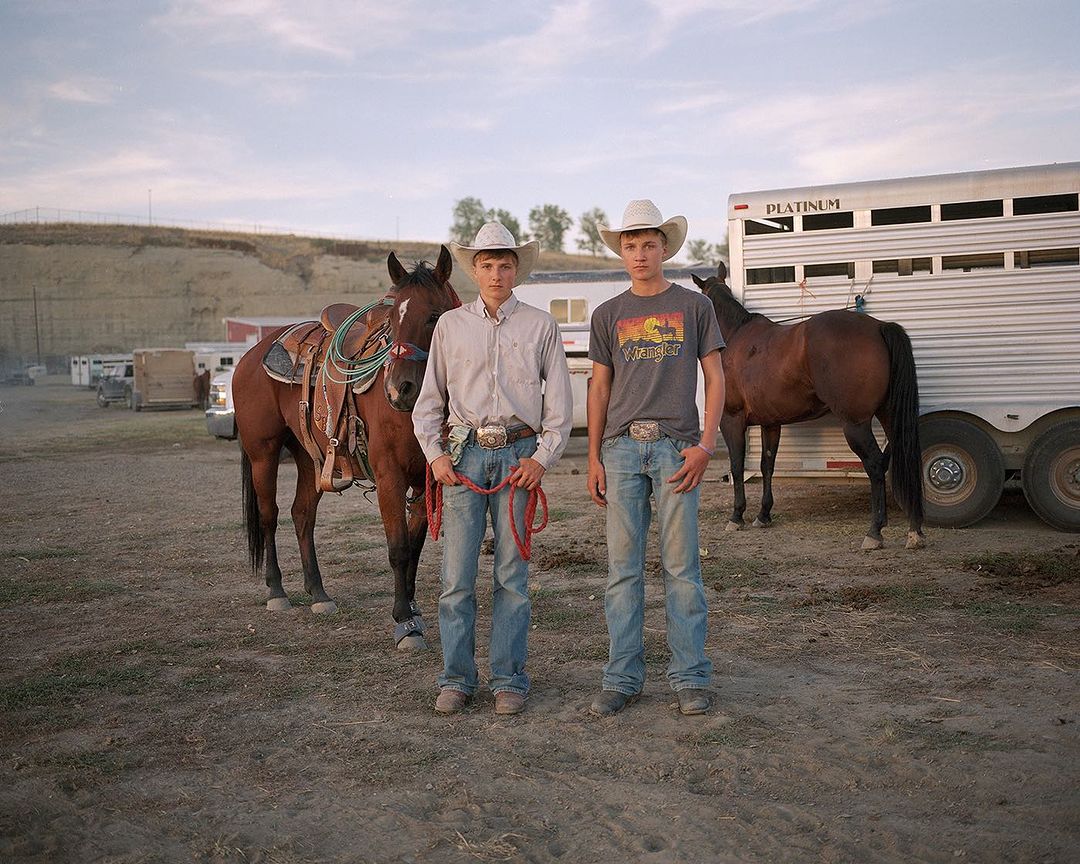 Two young cowboys with horses and a horse trailer.  Outdoor scene with dirt ground and overcast sky.  Casual western fashion style.