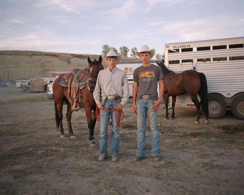 Two young cowboys with horses and a horse trailer.  Outdoor scene with dirt ground and overcast sky.  Casual western fashion style.