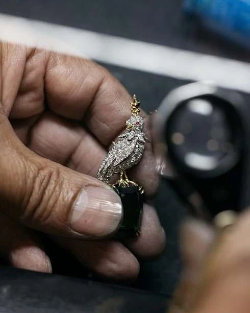 Close-up of a human hand holding an owl-shaped pendant, accented with diamonds and a dark green gemstone.