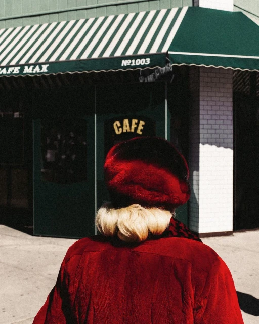 Woman in red coat and beret walks by 'Cafe Max' with green and white striped awning and a white brick facade on a sunny day.