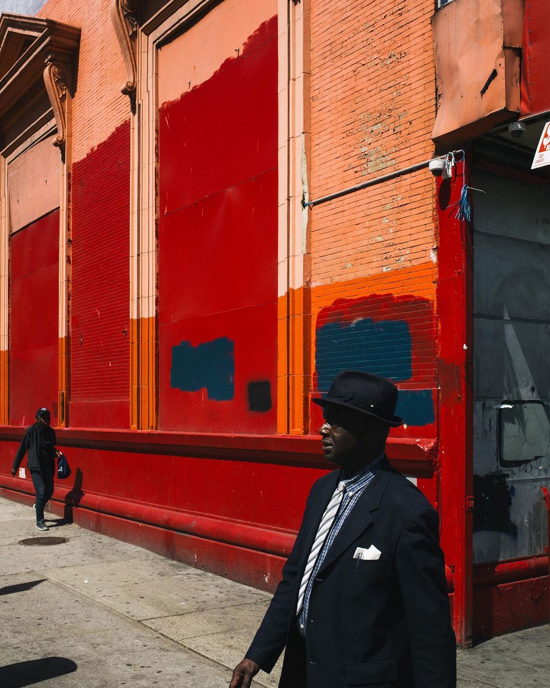 A dark-skinned adult male in a suit and hat walks past a red building on a city street, with another figure visible in the background.