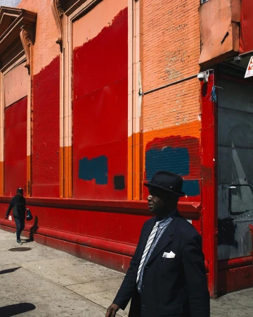 A dark-skinned adult male in a suit and hat walks past a red building on a city street, with another figure visible in the background.