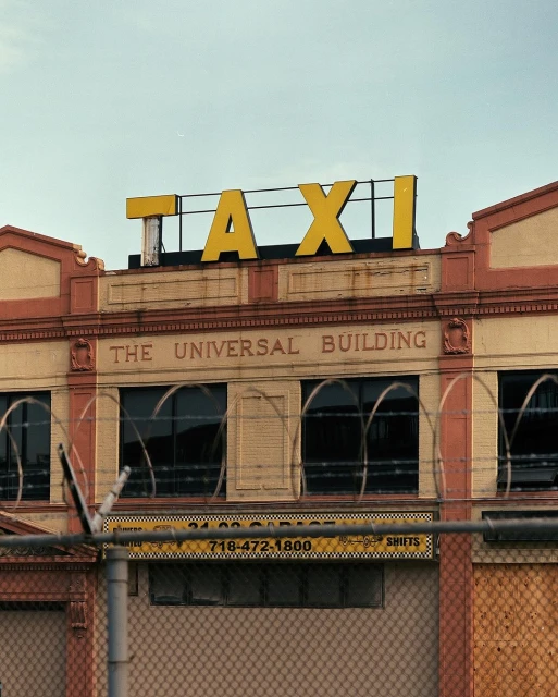 Exterior view of the Universal Building with a yellow 'TAXI' sign on top, brick facade, and a fence with barbed wire in the foreground.