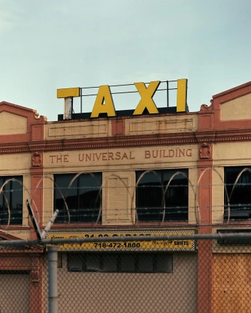 Exterior view of the Universal Building with a yellow 'TAXI' sign on top, brick facade, and a fence with barbed wire in the foreground.
