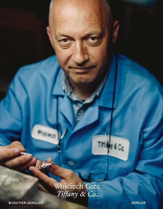 Close-up of a jeweler working on jewelry. Adult male, blue work shirt, indoor lighting, working with small tool. Tiffany & Co. logo visible.