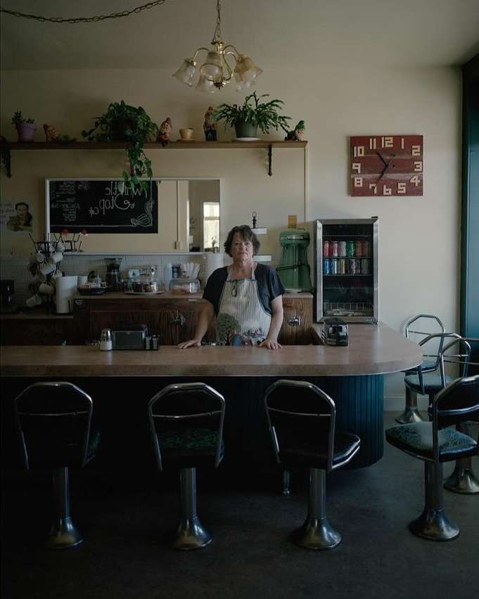 Inside a diner, a woman in an apron stands behind the counter with stools. A vintage clock and soda machine accent the scene.