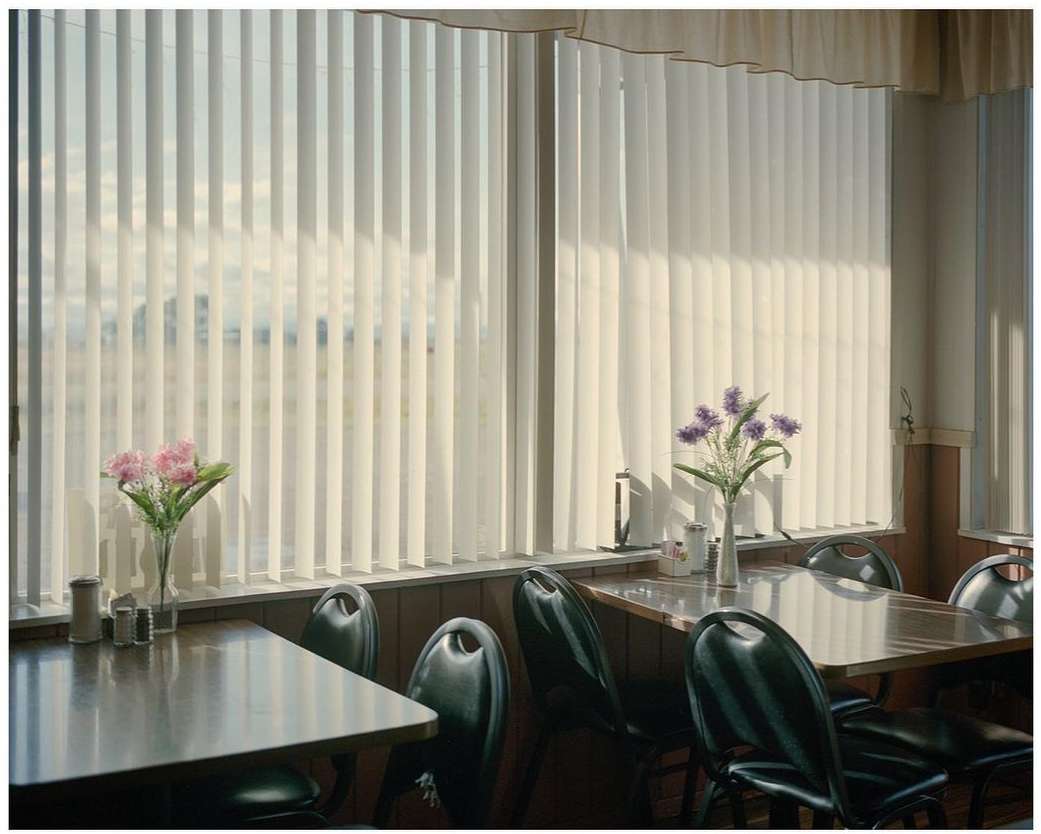 Diner interior featuring tables and chairs arranged near a window with blinds and floral arrangements casting soft light, creating a quiet ambiance.