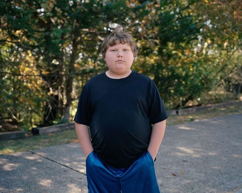 Portrait of a young person standing outdoors wearing a black t-shirt and blue athletic shorts, posing with hands in pockets.