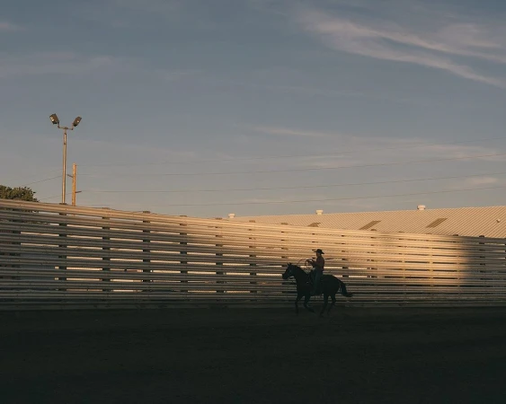A lone cowboy on horseback practices roping in a sunlit rodeo arena against a blue sky and wooden fence backdrop at golden hour.