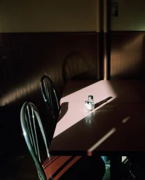 A diagonal shaft of sunlight illuminates salt and pepper shakers on a diner table with empty chairs.