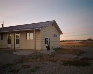 Exterior view of a pale yellow motel building with a standalone payphone against a dusk sky by Arnaud Montagard