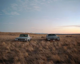 Two vintage cars parked in a dry grassy meadow under a pale sunset sky by photographer Arnaud Montagard