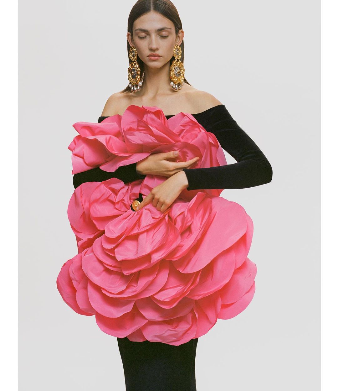 Editorial fashion shot of a woman in a black dress adorned with a large pink flower, accessorized with ornate gold earrings against a white backdrop.