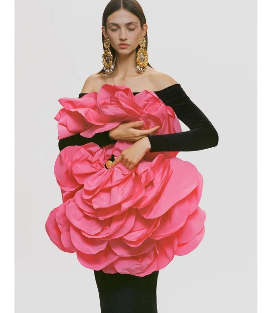 Editorial fashion shot of a woman in a black dress adorned with a large pink flower, accessorized with ornate gold earrings against a white backdrop.