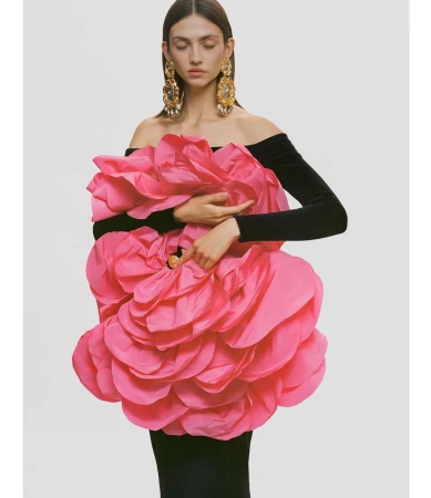 Editorial fashion shot of a woman in a black dress adorned with a large pink flower, accessorized with ornate gold earrings against a white backdrop.
