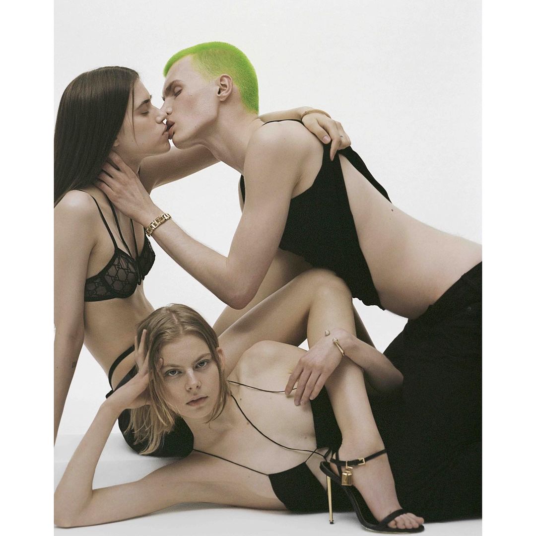 Fashion editorial photo features three young adults in black attire against a stark white backdrop, with one person kissing another.