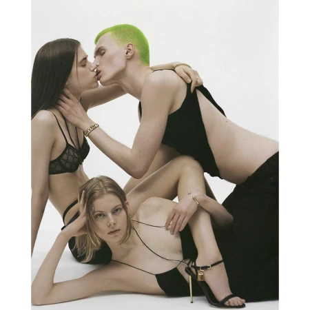 Fashion editorial photo features three young adults in black attire against a stark white backdrop, with one person kissing another.