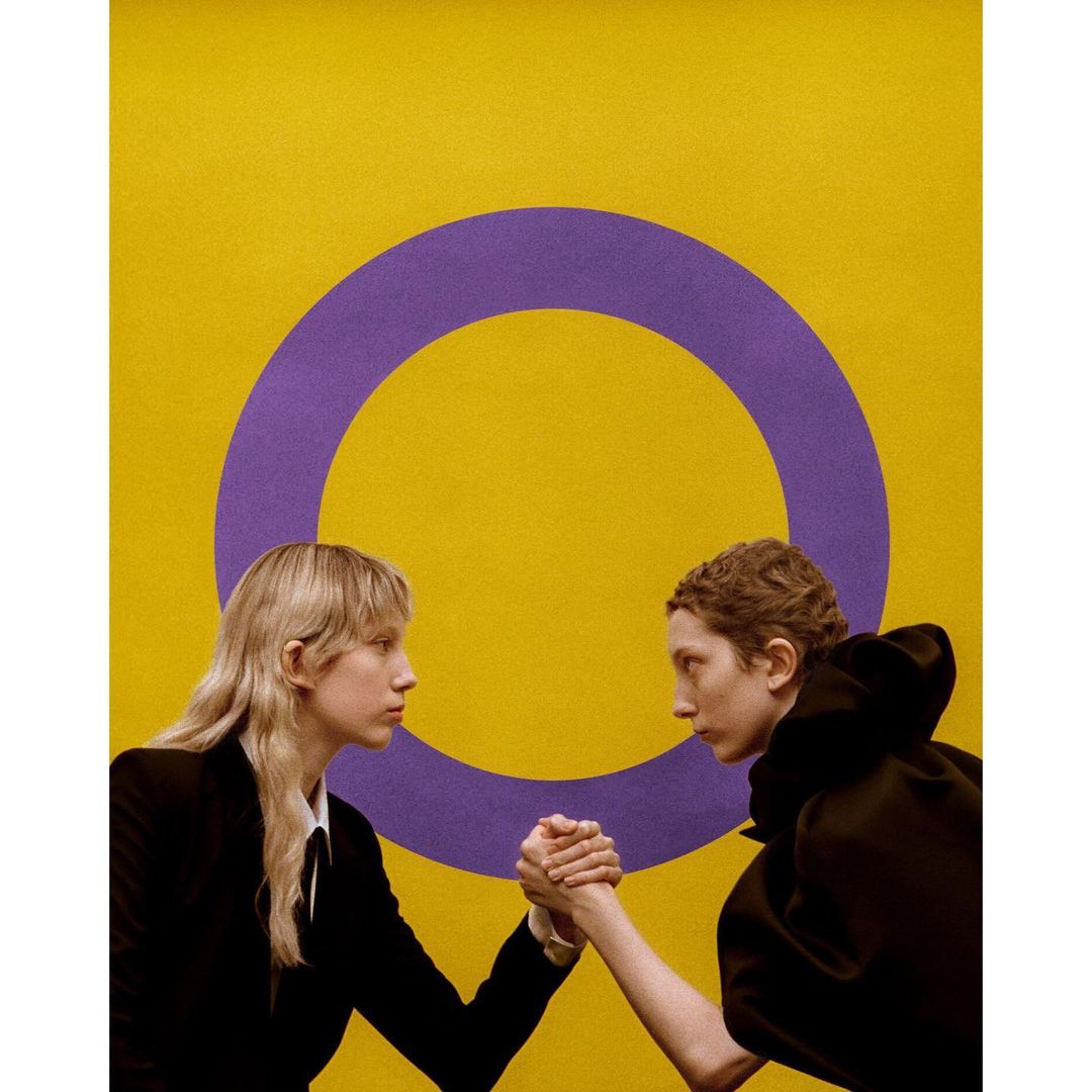 Two women arm wrestling against a yellow background with a purple circle, conveying competition and strength in a studio setting.