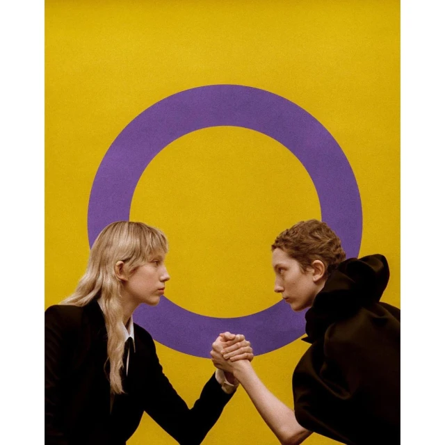Two women arm wrestling against a yellow background with a purple circle, conveying competition and strength in a studio setting.