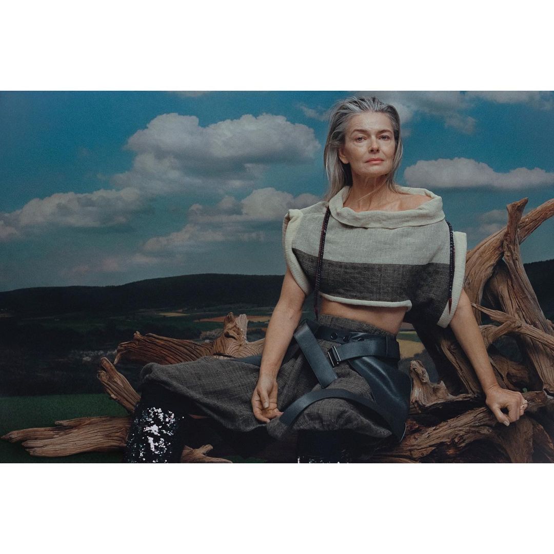 Silver-haired woman in a unique fashion ensemble sits on a fallen tree against a backdrop of green fields and cloudy blue skies.