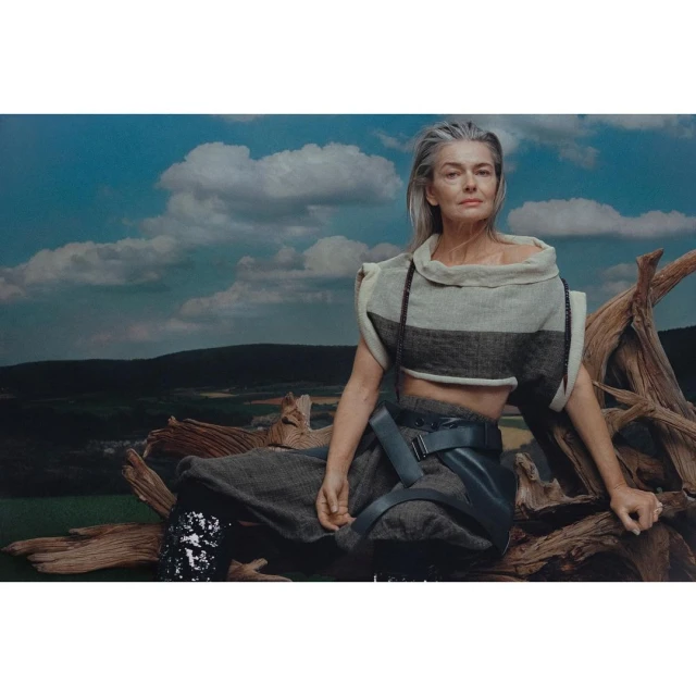 Silver-haired woman in a unique fashion ensemble sits on a fallen tree against a backdrop of green fields and cloudy blue skies.