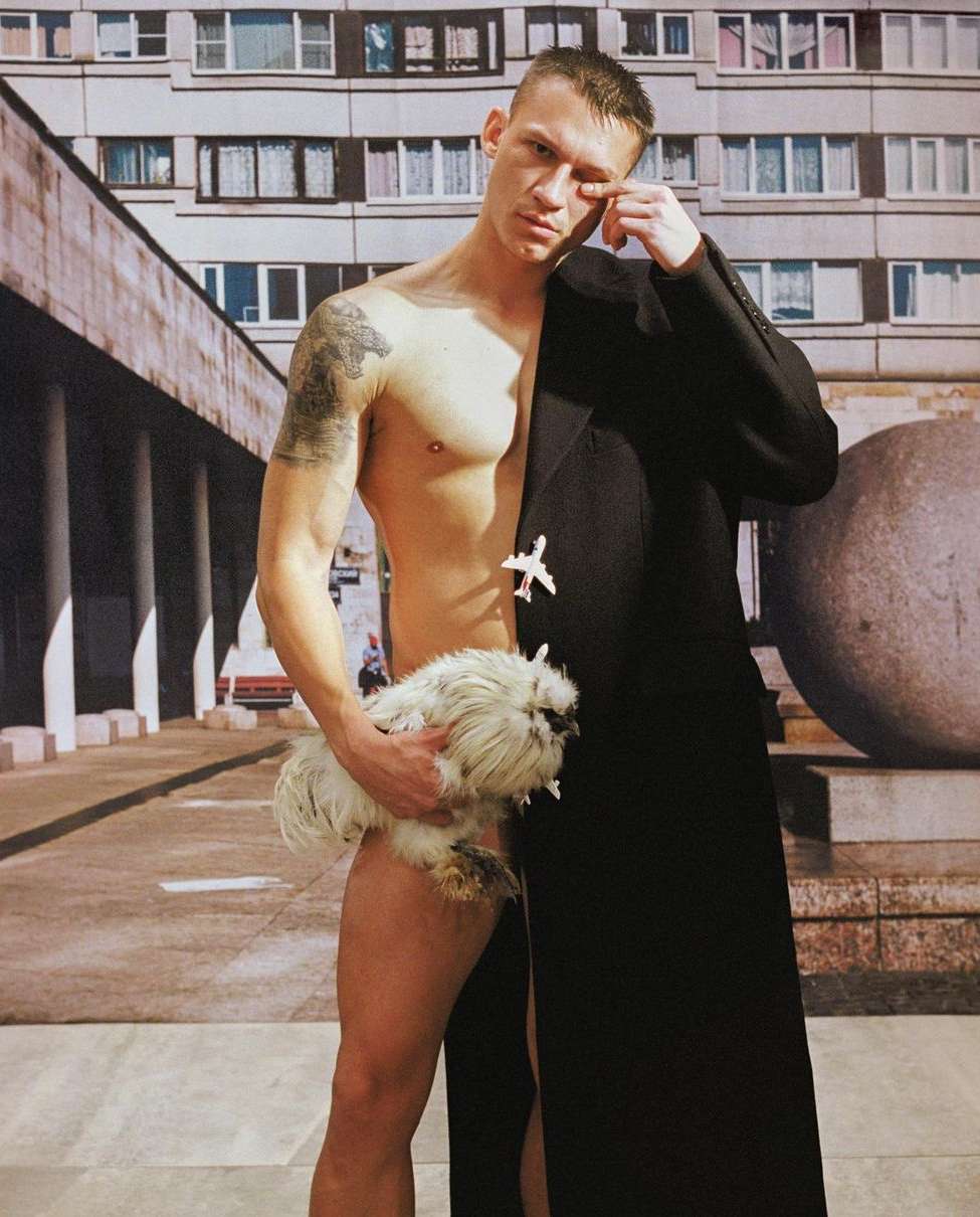 Man with bare torso holds a fluffy white chicken, stands partially in a black coat against a backdrop of a building and street.