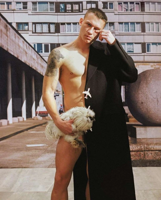 Man with bare torso holds a fluffy white chicken, stands partially in a black coat against a backdrop of a building and street.