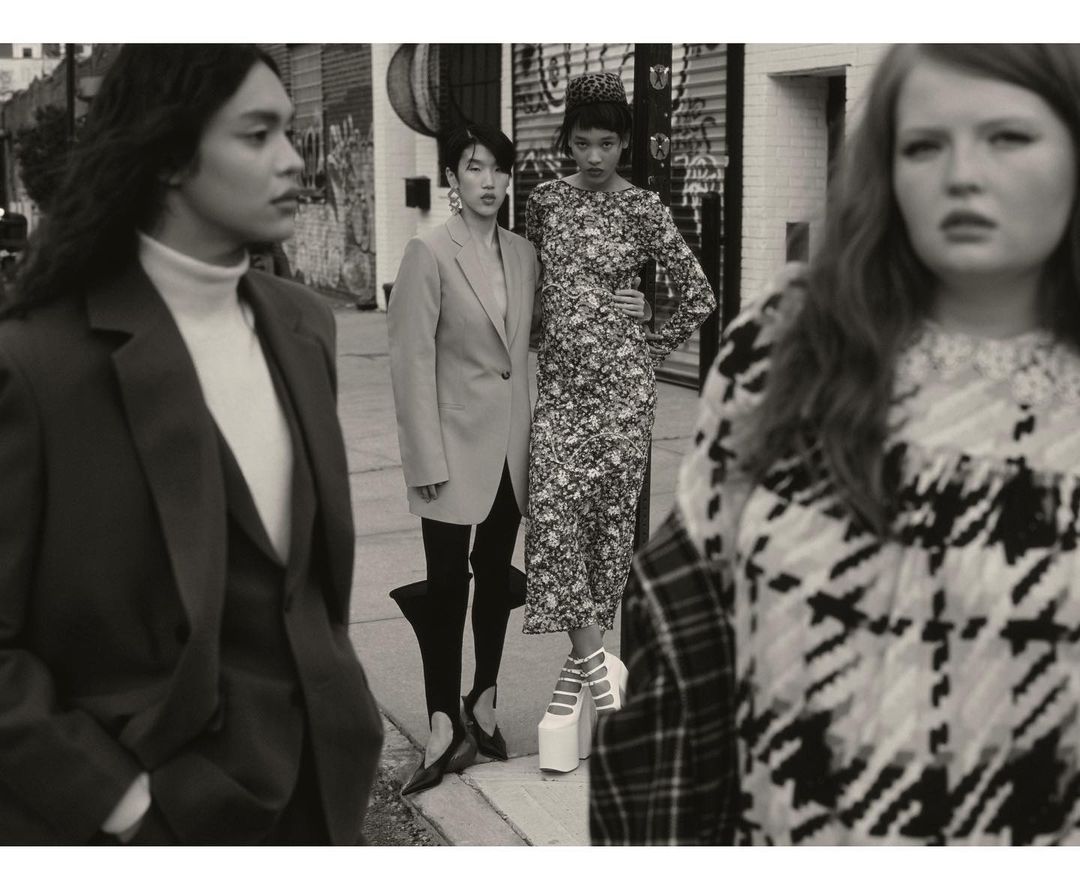 A fashion editorial shot of four diverse women in stylish outfits and platform heels, posing on a city street with building facades behind.