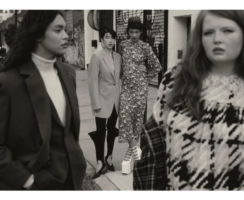 A fashion editorial shot of four diverse women in stylish outfits and platform heels, posing on a city street with building facades behind.