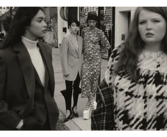 A fashion editorial shot of four diverse women in stylish outfits and platform heels, posing on a city street with building facades behind.