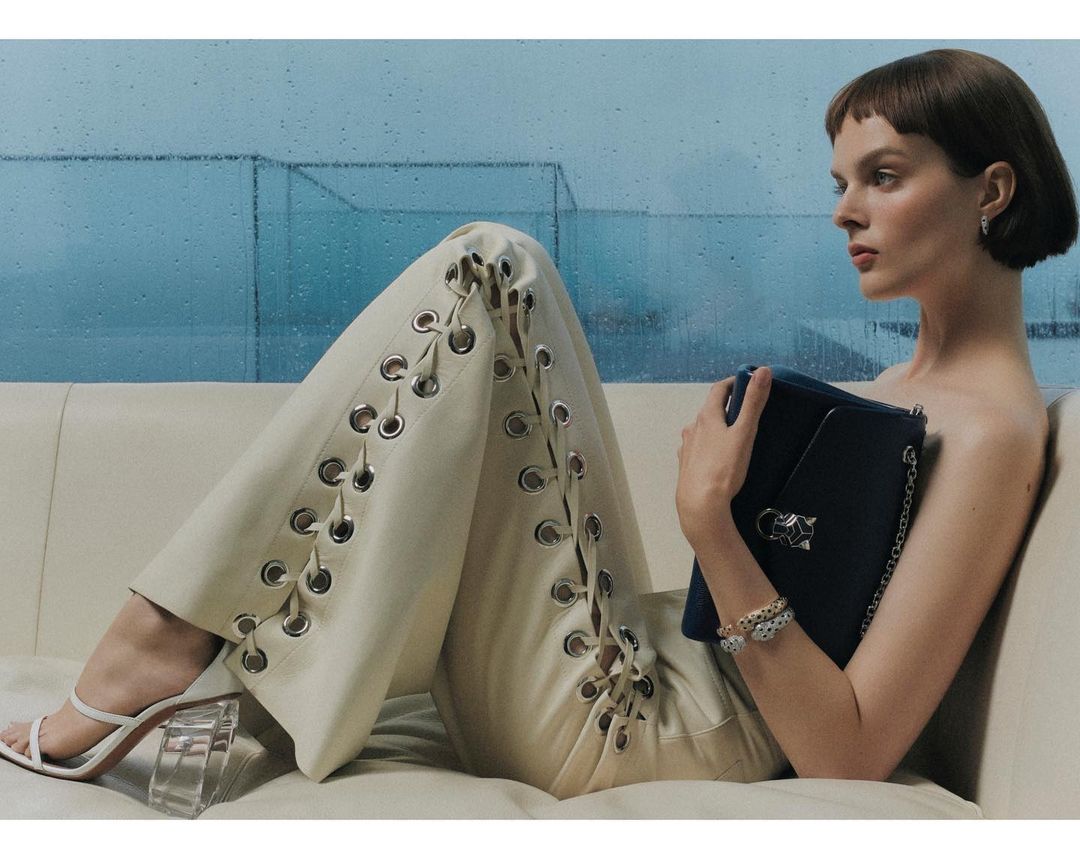 Elegant adult woman posing on a cream sofa with a blue clutch, wearing strappy heels and pants with grommet and lace-up detailing.