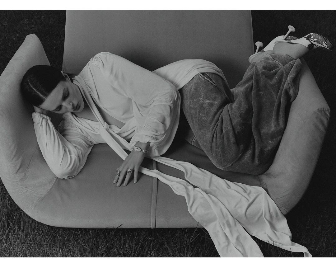 Black and white fashion shot of an adult woman reclining on a modern chair outdoors wearing a draped blouse and wide-leg velvet pants.