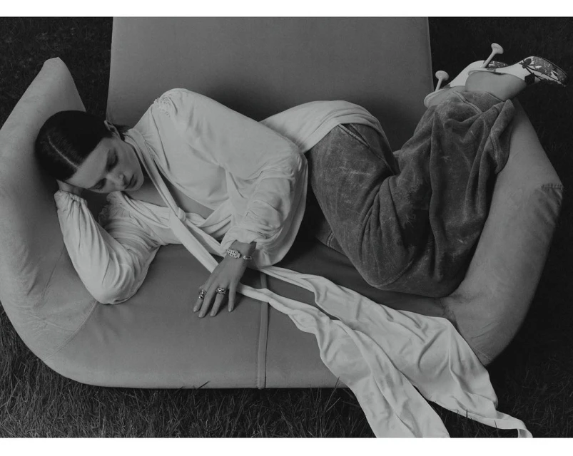 Black and white fashion shot of an adult woman reclining on a modern chair outdoors wearing a draped blouse and wide-leg velvet pants.