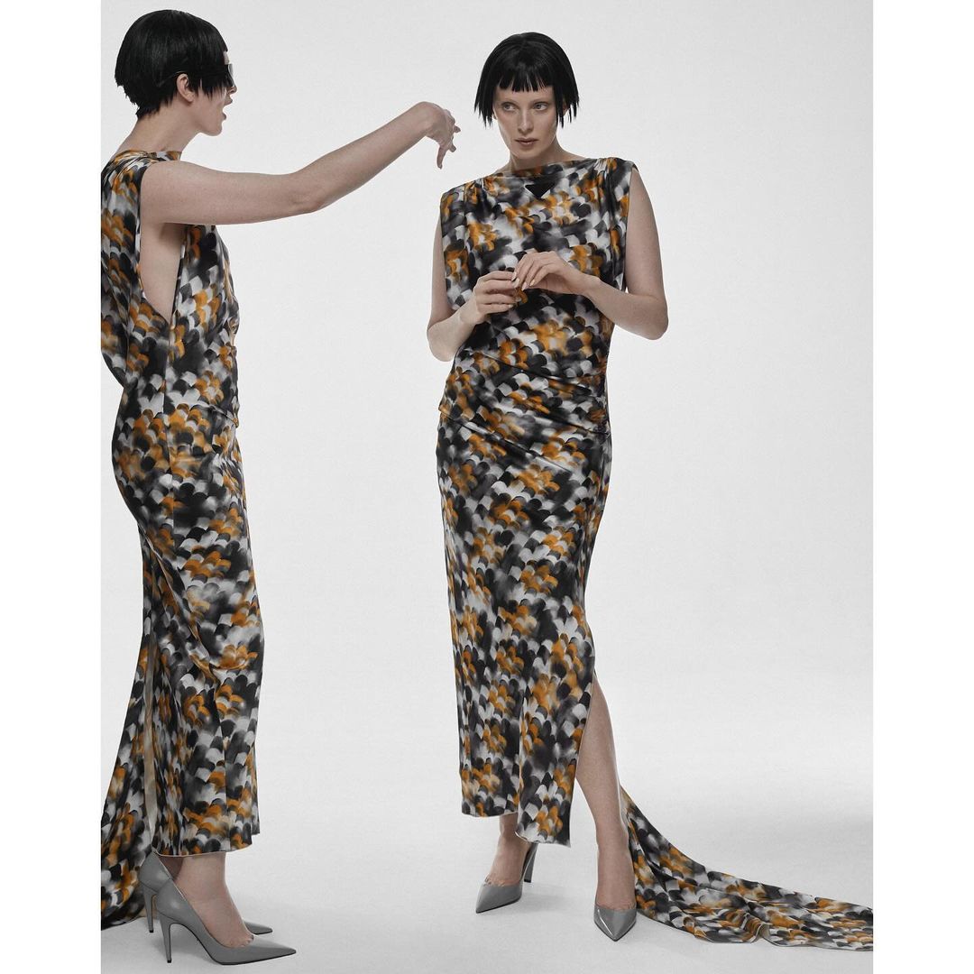 Two women pose in patterned dresses in a white studio. One model stands while the other reaches out, both wearing floor-length dresses.