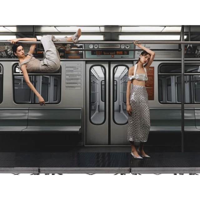 Two women in fashionable clothing inside a subway car, one hanging from a bar, the other standing, shot with studio lighting.