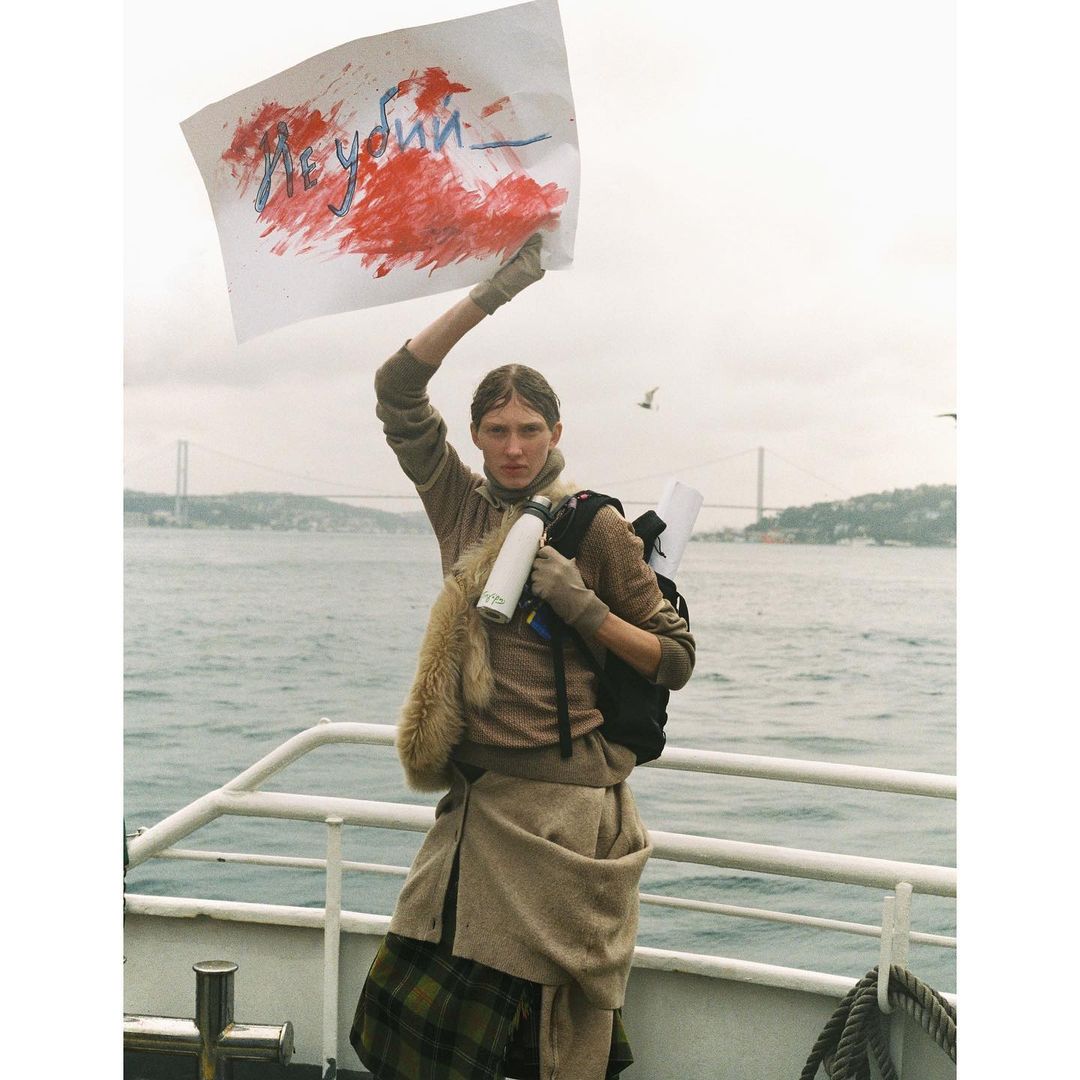 Adult female stands on a ferry holding a sign with painted text, wearing a tan coat, backpack and gloves against a sea background.