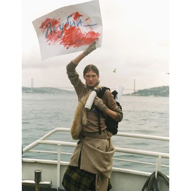 Adult female stands on a ferry holding a sign with painted text, wearing a tan coat, backpack and gloves against a sea background.