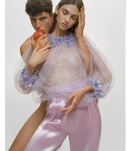 Fashion editorial shot of a topless man and a woman in a sheer lilac top and shiny lilac pants holding an apple against a white background.