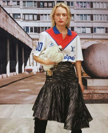 Model holding a fluffy chicken, wearing a polo shirt and dark flared skirt in front of a residential building exterior.