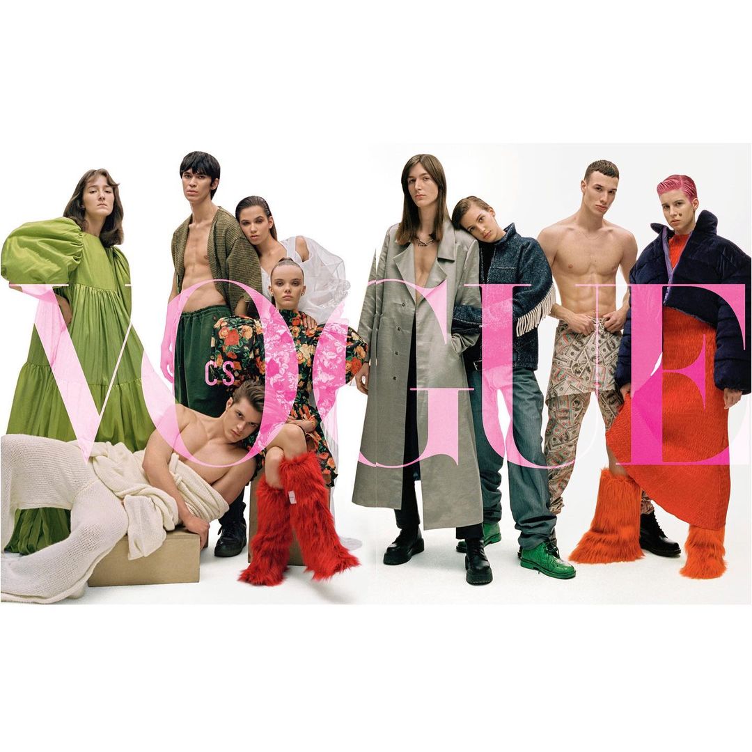 Group of eight fashion models in various outfits pose in front of a white backdrop, large pink Vogue text overlay. 